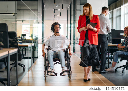 A business leader with her colleague, an African-American businessman who is a disabled person, pass by their colleagues who work in modern offices A business leader with her colleague, an African-American businessman who is a disabled person, pass by their colleagues who work in modern offices 114510571