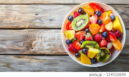 An inviting bowl of mixed fruit salad with vibrant colors, featuring strawberries, kiwi, oranges, and blueberries, on a rustic wooden background. Banner with copy space 114512650