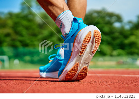 Athlete Wearing Bright Blue Running Shoes on Red Stadium Track During Training 114512823
