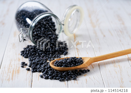 Raw black beans in wooden scoop and in glass storage jar on a white wooden table. Raw black beans in wooden scoop and in glass storage jar on a white wooden table. 114514218