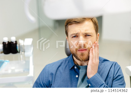 Sad man sitting in dental chair suffering from acute tooth pain, rubbing his cheek, close up 114514362