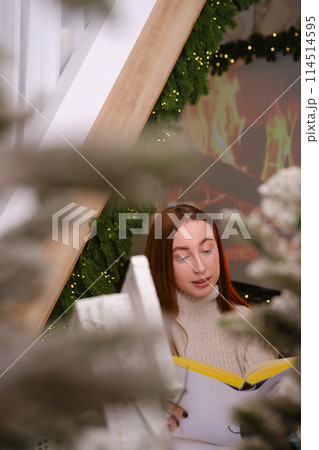 Hello winter. smiling modern woman in white sweater siting on chair and reading a book in room decorated for celebrating the new year and christmas festive mood 114514595