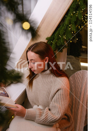 Hello winter. smiling modern woman in white sweater siting on chair and reading a book in room decorated for celebrating the new year and christmas festive mood 114514596