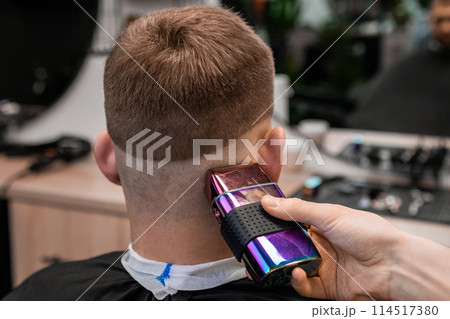 Hairstylist removes hair behind client ear and occiput with shaver in barbershop closeup. Barber uses modern machine for stylish haircut in salon Hairstylist removes hair behind client ear and occiput with shaver in barbershop closeup. Barber uses modern machine for stylish haircut in salon 114517380