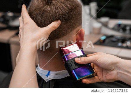 Barber uses shaver machine to cut young man hair in barbershop closeup. Master does modern haircut to male client in professional salon Barber uses shaver machine to cut young man hair in barbershop closeup. Master does modern haircut to male client in professional salon 114517388