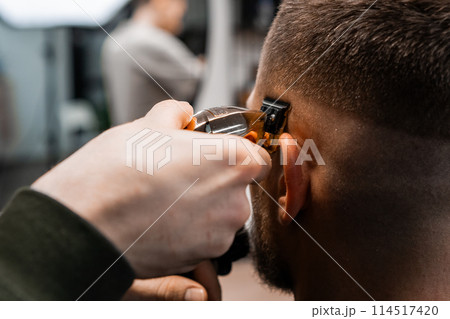 Skilled barber uses automatic trimmer to cut client hair on temple in barbershop closeup. Hairdresser does stylish hairdo by salon mirror Skilled barber uses automatic trimmer to cut client hair on temple in barbershop closeup. Hairdresser does stylish hairdo by salon mirror 114517420