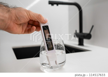 Man hand immerses conductometer in a beaker of water to check purity in front of a modern kitchen water faucet. Conductometer shows that water is dirty Man hand immerses conductometer in a beaker of water to check purity in front of a modern kitchen water faucet. Conductometer shows that water is dirty 114517633