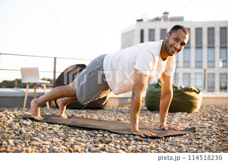 Strong male in plank position on mat during sunrise against urban background. Strong male in plank position on mat during sunrise against urban background. 114518230