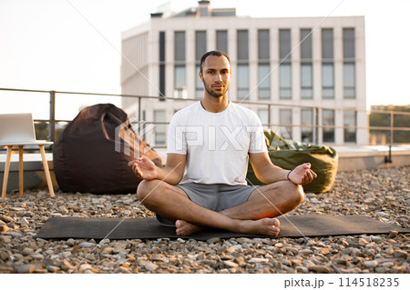 Male meditating in lotus position while sitting on mat on roof of modern house. 114518235