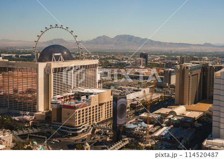 Aerial view of Las Vegas Strip in daytime with clear sky, sun shining on cityscape. High Roller observation wheel and city buildings stand out. Vibrant urban environment captured. 114520487