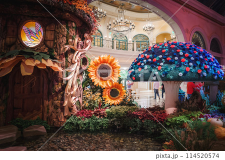 Vibrant indoor scene in Las Vegas Strip hotel with whimsical tree, colorful floral mushroom, and lush garden. Luxurious decor and lively visitors add to enchanting ambiance. 114520574
