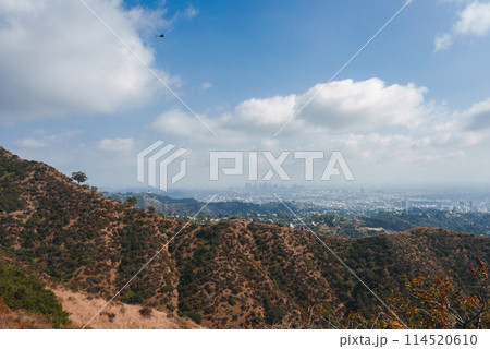 Scenic view of arid hills surrounding Los Angeles with a cityscape in the distance under a partly cloudy sky. Helicopter adds dynamism to the tranquil scene. 114520610