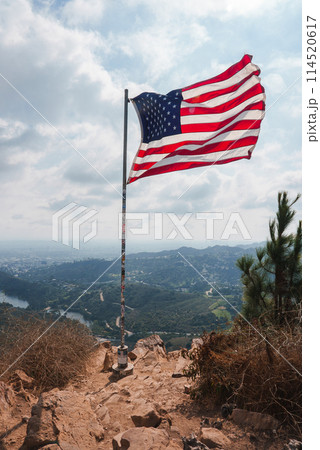 American flag on flagpole waving against hilly backdrop. Rugged terrain leads to flagpole, cloudy sky with natural light. View reminiscent of Los Angeles hills. 114520617