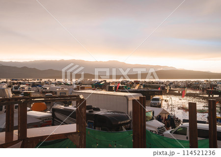 Desert music and art festival scene from an elevated view at either sunrise or sunset. Vehicles and creative structures against a backdrop of a desert landscape. 114521236