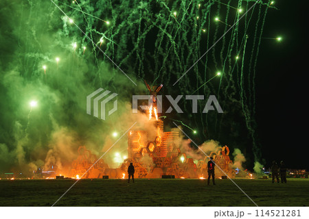 Vibrant night scene at desert music and art festival spectators silhouetted against bright lights, fiery structure engulfed in flames, green fireworks in sky. Typical of desert festivals. 114521281
