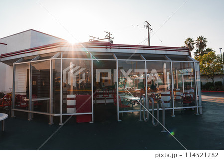 Classic American diner in Los Angeles with mid 20th century style, white exterior, red trim, traditional booth seating, and outdoor area with red and white chairs under a canopy. 114521282
