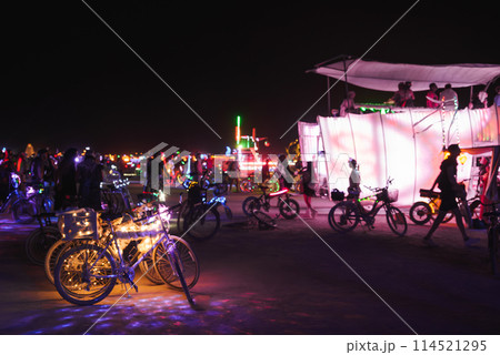 Vibrant night scene at a music and art festival in the desert. Colorful lights illuminate the atmosphere. Festive attendees on light adorned bicycles. Vibrant night scene at a music and art festival in the desert. Colorful lights illuminate the atmosphere. Festive attendees on light adorned bicycles. 114521295