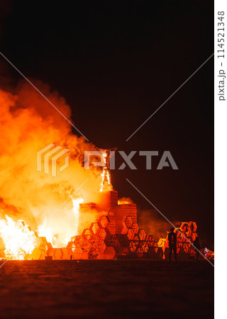 Experience the mesmerizing glow of a massive bonfire illuminating a desert festival at night. Attendees observe a flaming structure in a communal setting. Experience the mesmerizing glow of a massive bonfire illuminating a desert festival at night. Attendees observe a flaming structure in a communal setting. 114521348