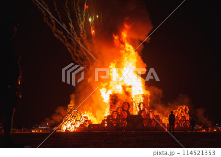 Mesmerizing image of intricate structure burning in desert at night. Spectacular flames, fireworks, and silhouetted spectators create dramatic scene. Symbolizes music and art festival. 114521353