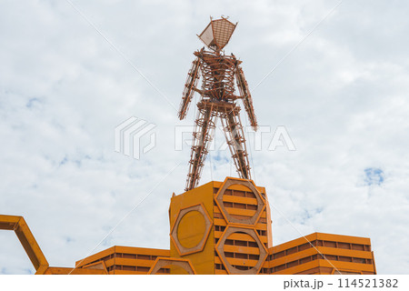 Intricate wooden figure with outstretched arms atop geometric base in vibrant orange hue. Possible art installation at desert festival, reflecting creativity and communal expression. 114521382