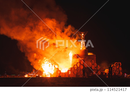 Dramatic night scene with burning wooden structure at desert art festival. Symbolizes impermanence, creativity, and community. Lights and people in background. 114521417