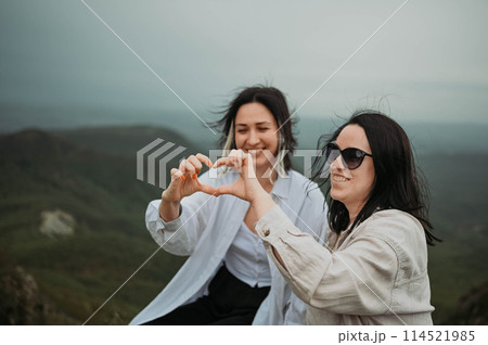 Young beautiful happy lesbian couple posing in cloudy mountains showing love sign outside at nature summer park. LGBT community concept. Female friends smiling enjoying moments together 114521985