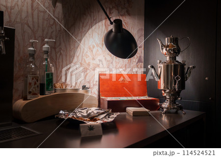 Luxury hotel countertop in Zermatt ski resort, Switzerland. Dark material contrasts with transparent bottles, white cups, orange box, and silver samovar. Modern lamp provides warm light ambiance. 114524521