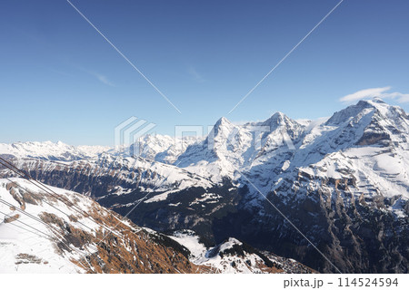Breathtaking panoramic view of snow covered Swiss Alps from Murren ski resort. Majestic mountains, ski lift, village below, tranquil skies. Ideal for winter activities. 114524594