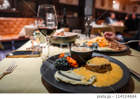 Dining scene with gourmet steak and vegetables on plate, glass of red wine, and cozy ambiance. Smartphone and fork on table. Other diners in background. Dining scene with gourmet steak and vegetables on plate, glass of red wine, and cozy ambiance. Smartphone and fork on table. Other diners in background. 114524684
