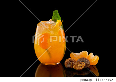 Closeup glass of orange tangerine fresh juice with ice cubes and a group of mandarin slices and figs isolated at black background. Closeup glass of orange tangerine fresh juice with ice cubes and a group of mandarin slices and figs isolated at black background. 114527452