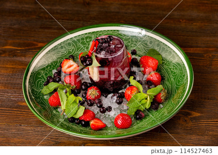 Black currant cocktail with berries and frappe at decorated with strawberry and mint plate at wooden table background. 114527463
