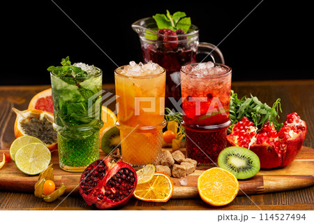 Three glasses of homemade healthy lemonades with tarragon, mint, citrus mix nd pomegranate at wooden table decorated with fruits background. 114527494