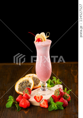 Glass of fresh strawberry milkshake decorated with cocktail cherry, banana slices and mint at wooden table isolated at black background. Glass of fresh strawberry milkshake decorated with cocktail cherry, banana slices and mint at wooden table isolated at black background. 114527527