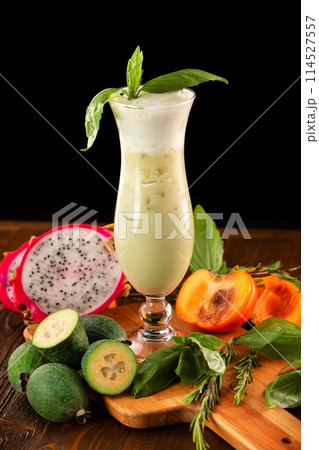 Glass of fresh fruit smoothie decorated with basil, feijoa, persimmon and dragon fruit at wooden table isolated at black background. Glass of fresh fruit smoothie decorated with basil, feijoa, persimmon and dragon fruit at wooden table isolated at black background. 114527557