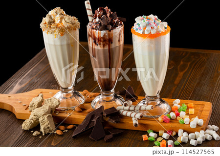 Three sweet milkshakes with chocolate, marshmallow, halva and whipped cream at a wooden board on a table isolated at black background. Three sweet milkshakes with chocolate, marshmallow, halva and whipped cream at a wooden board on a table isolated at black background. 114527565
