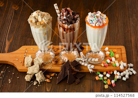 Three sweet milkshakes with chocolate, marshmallow, halva and whipped cream at a wooden board on table background. 114527568