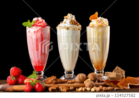 Three sweet milkshakes with nuts, caramel, strawberry and whipped cream at a wooden board on table background. Three sweet milkshakes with nuts, caramel, strawberry and whipped cream at a wooden board on table background. 114527582