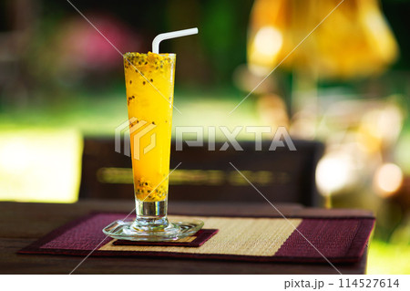 Closeup glass of passion fruit shake served with a straw at a wooden table at tropical outdoors background. Closeup glass of passion fruit shake served with a straw at a wooden table at tropical outdoors background. 114527614