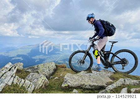 Cyclist man riding electric mountain bike outdoors. Male tourist biking along grassy trail in the mountains, wearing helmet and backpack. Concept of sport, active leisure and nature. Cyclist man riding electric mountain bike outdoors. Male tourist biking along grassy trail in the mountains, wearing helmet and backpack. Concept of sport, active leisure and nature. 114529921