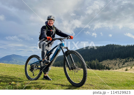 Cyclist man riding electric bike outdoors. Portrait of smiling male tourist resting on grassy hill, enjoying beautiful mountain landscape, wearing helmet. Concept of active leisure. 114529923