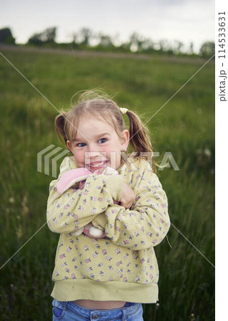 a beautiful blond girl with her toy bunny is playing in the field 114533631