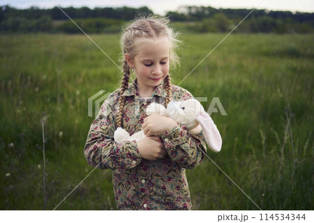 a beautiful blond girl with her toy bunny is playing in the field 114534344