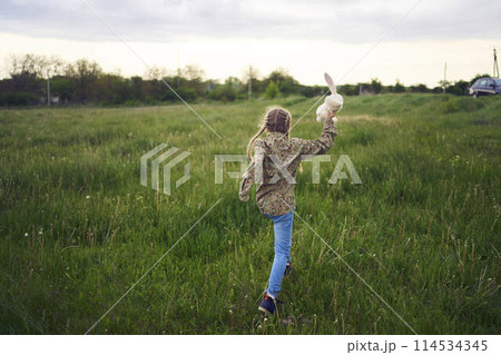 a beautiful blond girl with her toy bunny is playing in the field 114534345