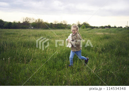 a beautiful blond girl with her toy bunny is playing in the field 114534348