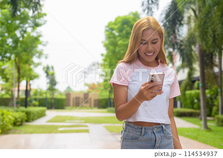 Portrait of beautiful Asian girl with blonde hair smiling using phone outdoors 114534937