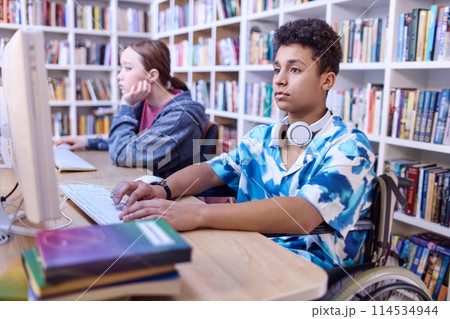 Portrait of teen boy using wheelchair in computer class at school library and doing research for project 114534944