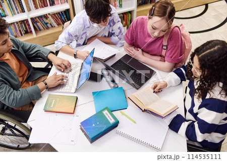 High angle view of diverse group of students sitting at table in school library together and doing research with textbooks and computers copy space High angle view of diverse group of students sitting at table in school library together and doing research with textbooks and computers copy space 114535111
