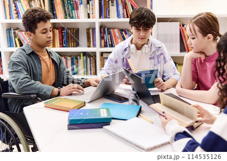 Inclusive group of students doing homework together sitting at table in school library and holding books 114535126