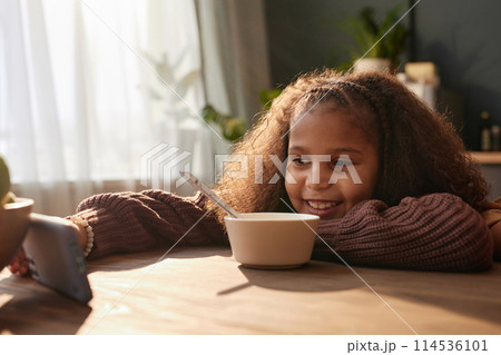 Portrait of smiling Black girl using smartphone at kitchen table in morning scene lit by cozy sunlight copy space Portrait of smiling Black girl using smartphone at kitchen table in morning scene lit by cozy sunlight copy space 114536101
