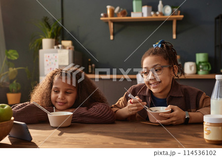 Front view portrait of two young Black girls eating breakfast in cozy kitchen lit by sunlight Front view portrait of two young Black girls eating breakfast in cozy kitchen lit by sunlight 114536102